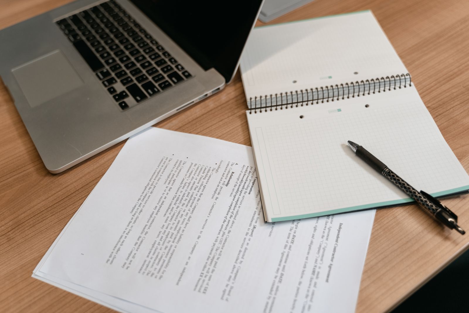 Legal paperwork, an open notebook and a pen arranged beside a laptop on a wooden desk.