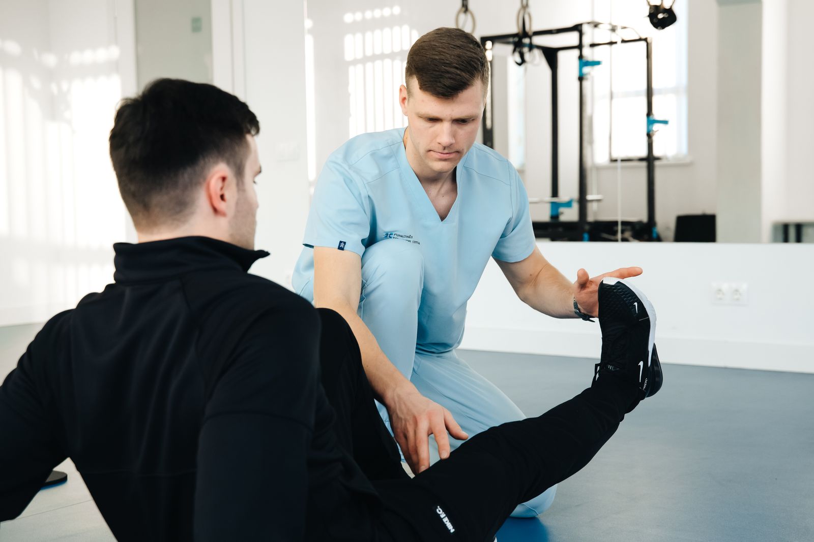 A physiotherapist in clinical scrubs supports a patient during a leg rehabilitation exercise.